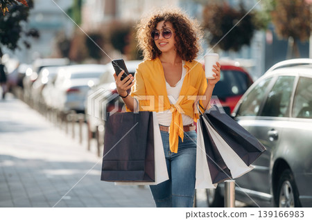 Cup of drink and smartphone in hands. Happy woman is with shopping bags in town 139166983