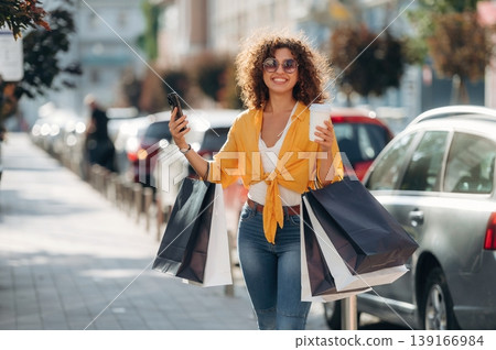 Cup of drink and smartphone in hands. Happy woman is with shopping bags in town Cup of drink and smartphone in hands. Happy woman is with shopping bags in town 139166984