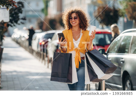 Cup of drink and smartphone in hands. Happy woman is with shopping bags in town Cup of drink and smartphone in hands. Happy woman is with shopping bags in town 139166985