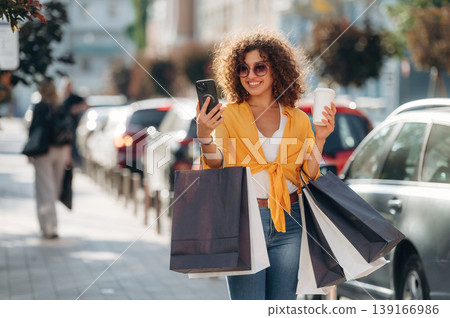 Cup of drink and smartphone in hands. Happy woman is with shopping bags in town Cup of drink and smartphone in hands. Happy woman is with shopping bags in town 139166986