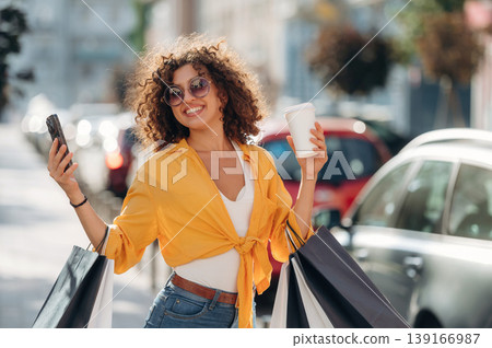 Cup of drink and smartphone in hands. Happy woman is with shopping bags in town Cup of drink and smartphone in hands. Happy woman is with shopping bags in town 139166987