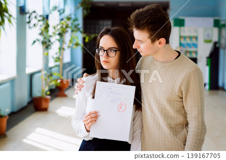 Young attractive student girl standing in corridor of campus with result of test looks aside, receives F grade for her work. Her classmate and good friend tries to calm her down and help in studying 139167705