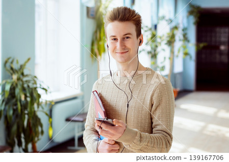 Happy student with mobile phone, earphones in his ears and books in his hands looking to the camera. Young cheerful boy in university campus using modern technologies 139167706