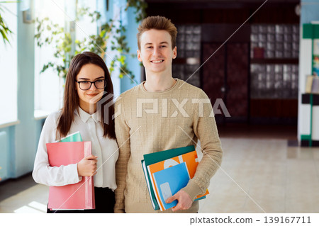 Portrait of two high-school learners with course books, text-books and folders before classes in school corridor in sunny day. Cheerful students are eager to begin new education year Portrait of two high-school learners with course books, text-books and folders before classes in school corridor in sunny day. Cheerful students are eager to begin new education year 139167711
