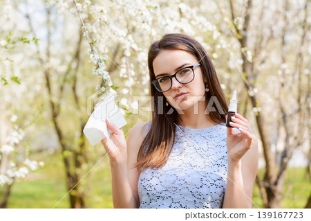 Girl holding respiratory nasal spray and white handkerchief against spring bloom in park. Portrait of a woman having allergy on flower pollen Girl holding respiratory nasal spray and white handkerchief against spring bloom in park. Portrait of a woman having allergy on flower pollen 139167723