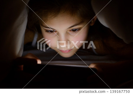Beautiful caucasian girl playing games on her mobile phone under the blanket at night. Little child don`t want to sleep and plays on smartphone under the cover in order parents don`t see her 139167750