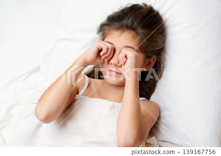Sleepy pretty little child rubs her eyes in the morning. Lovely small child with long dark natural hair lies on comfortable bed rubbing eyes and yawning 139167756