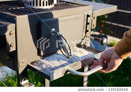 Hand pulls an ash tray filled with ash from outdoor grill. Concept of barbecue cleaning Hand pulls an ash tray filled with ash from outdoor grill. Concept of barbecue cleaning 139168408
