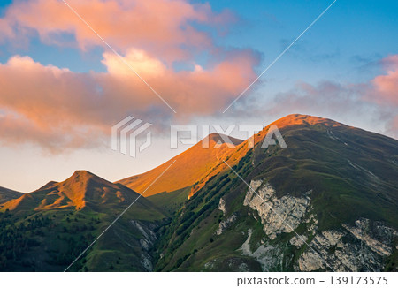 Mountain peaks bathed in warm golden light under a sky with soft orange clouds at an early morning or late evening Mountain peaks bathed in warm golden light under a sky with soft orange clouds at an early morning or late evening 139173575
