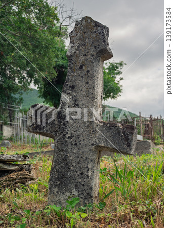 Ancient stone cross monument rises from overgrown grass in a quiet rural cemetery on a cloudy afternoon Ancient stone cross monument rises from overgrown grass in a quiet rural cemetery on a cloudy afternoon 139173584