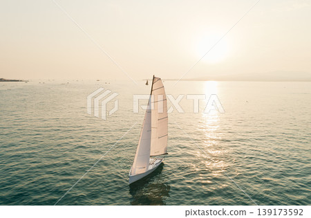 Aerial view captures solitary white sailboat gracefully navigates tranquil waters reflecting the warm glow of a beautiful sunset Aerial view captures solitary white sailboat gracefully navigates tranquil waters reflecting the warm glow of a beautiful sunset 139173592
