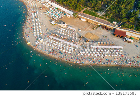 Aerial view shows a vibrant, crowded summer beach with numerous swimmers in the clear blue sea and sunbathers lining the shore Aerial view shows a vibrant, crowded summer beach with numerous swimmers in the clear blue sea and sunbathers lining the shore 139173595