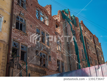 Damaged urban brick building with shattered windows and crumbling walls featuring green safety netting under blue sky Damaged urban brick building with shattered windows and crumbling walls featuring green safety netting under blue sky 139173647