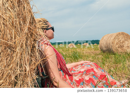 Woman with blonde dreadlocks relaxes by a large hay bale in a sunny summer field, enjoying the peaceful farm scenery Woman with blonde dreadlocks relaxes by a large hay bale in a sunny summer field, enjoying the peaceful farm scenery 139173658
