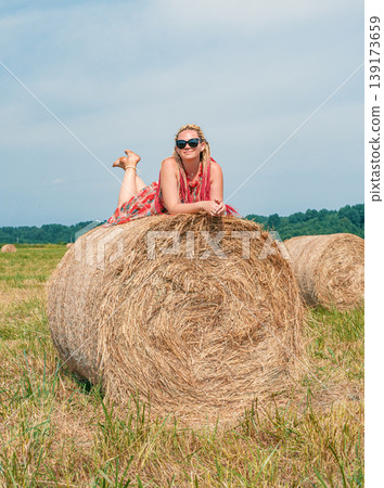 A smiling woman with blonde dreadlocks and sunglasses lies on a large hay bale in a vast, sunny rural field. A smiling woman with blonde dreadlocks and sunglasses lies on a large hay bale in a vast, sunny rural field. 139173659