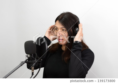 Confident LGBTQ Asian woman wearing glasses adjusting headphones while recording voice with microphone in studio minimal background modern content creator lifestyle focused expression Confident LGBTQ Asian woman wearing glasses adjusting headphones while recording voice with microphone in studio minimal background modern content creator lifestyle focused expression 139173800