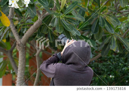 Person wearing hoodie using camera with telephoto lens photographing flowers in green garden outdoor nature hobby creative photography activity with focus on plants Person wearing hoodie using camera with telephoto lens photographing flowers in green garden outdoor nature hobby creative photography activity with focus on plants 139173816