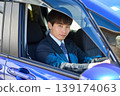 A young Japanese businessman looks at the camera through the window from the driver's seat of his company car. 139174063