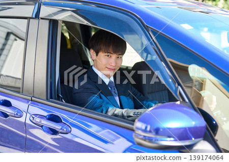 A young Japanese businessman looks at the camera through the window from the driver's seat of his company car. A young Japanese businessman looks at the camera through the window from the driver's seat of his company car. 139174064