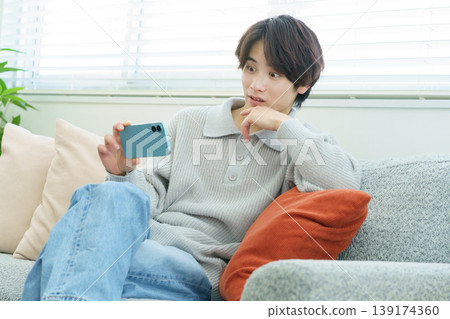 A Japanese man with a surprised look on his face is watching a video on his smartphone in his living room. A Japanese man with a surprised look on his face is watching a video on his smartphone in his living room. 139174360