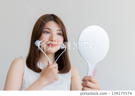 A woman using a facial roller for self-care while looking in a hand mirror – an image of beauty and lifting. 139175574