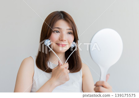 A woman using a facial roller for self-care while looking in a hand mirror – an image of beauty and lifting. 139175575