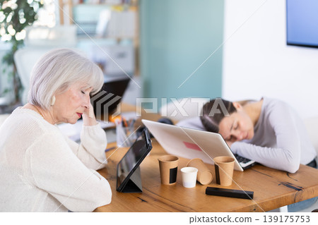 Tired elderly and adult female colleagues fall asleep while working Tired elderly and adult female colleagues fall asleep while working 139175753
