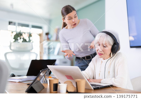 Adult woman scolding female colleague with headphones at laptop Adult woman scolding female colleague with headphones at laptop 139175771