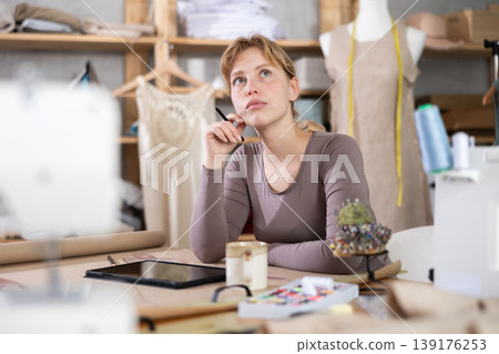 Young sad female dressmaker sitting at the table in sewing workshop Young sad female dressmaker sitting at the table in sewing workshop 139176253