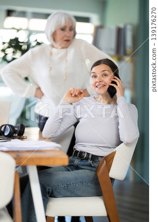 Elderly woman judging female colleague talking on mobile phone Elderly woman judging female colleague talking on mobile phone 139176270