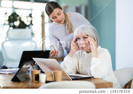 Adult woman sympathizes to sick senior female manager with headache and offers pain pills. Adult woman sympathizes to sick senior female manager with headache and offers pain pills. 139176359