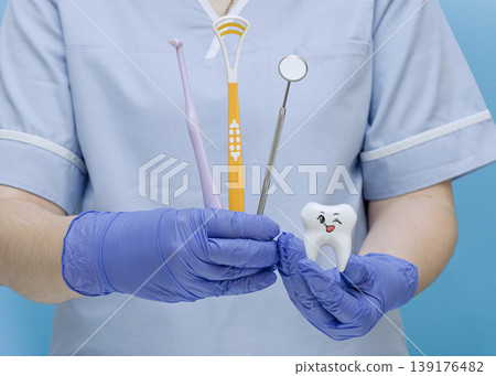 Friendly dentist in blue gloves showing a mouth mirror, tongue scraper, brush, and a happy tooth figurine. Friendly dentist in blue gloves showing a mouth mirror, tongue scraper, brush, and a happy tooth figurine. 139176482
