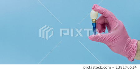 Dentist hand in a pink medical glove holding a dental implant on a blue background with copy space. Dentist hand in a pink medical glove holding a dental implant on a blue background with copy space. 139176514