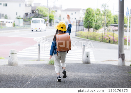 A back view of an elementary school student with a schoolbag on their back, raising their hand as they cross a road at a crosswalk; a faceless image representing traffic safety for going to and from school. A back view of an elementary school student with a schoolbag on their back, raising their hand as they cross a road at a crosswalk; a faceless image representing traffic safety for going to and from school. 139176875