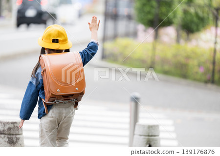 A back view of an elementary school student with a schoolbag on their back, raising their hand as they cross a road at a crosswalk; a faceless image representing traffic safety for going to and from school. A back view of an elementary school student with a schoolbag on their back, raising their hand as they cross a road at a crosswalk; a faceless image representing traffic safety for going to and from school. 139176876