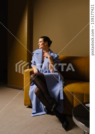 Model sits on a couch wearing blue coat and black stockings in modern room during daytime Model sits on a couch wearing blue coat and black stockings in modern room during daytime 139177421