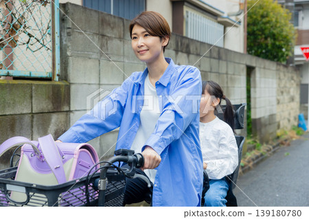 A woman riding a bicycle with an elementary school child on the back. A woman riding a bicycle with an elementary school child on the back. 139180780
