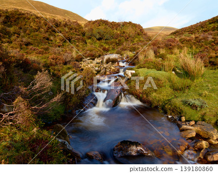 Mountain stream cascades down rocky slope through green and brown heather in daylight. Mountain stream cascades down rocky slope through green and brown heather in daylight. 139180860