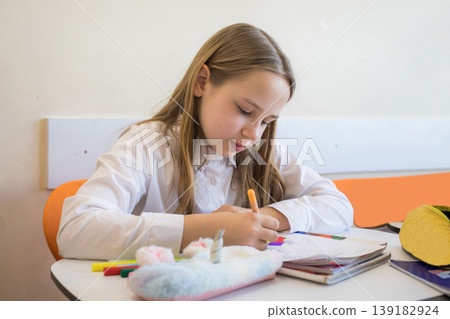 Happy young girl student sitting at desk writing in notebook in school classroom. Happy young girl student sitting at desk writing in notebook in school classroom. 139182924