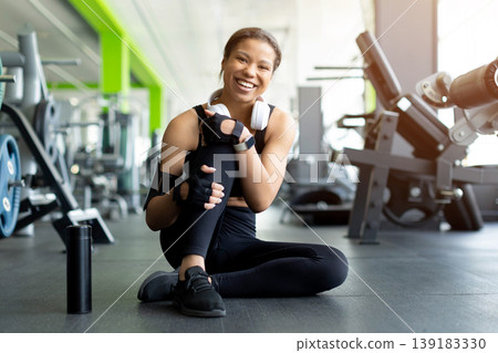 Happy Black woman sitting on gym floor hugging her knee and laughing Happy Black woman sitting on gym floor hugging her knee and laughing 139183330