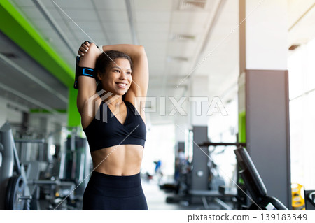 Smiling Black woman in black sports bra stretching arms overhead in gym Smiling Black woman in black sports bra stretching arms overhead in gym 139183349
