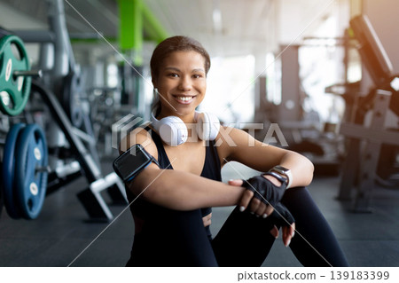 Happy young Black woman sitting on gym floor resting after workout Happy young Black woman sitting on gym floor resting after workout 139183399