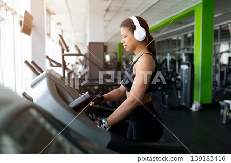 Young Black woman walking on treadmill in gym, wearing white headphones Young Black woman walking on treadmill in gym, wearing white headphones 139183416
