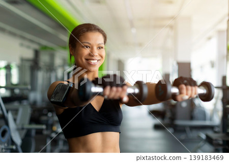 Smiling young Black woman lifting two dumbbells forward in gym Smiling young Black woman lifting two dumbbells forward in gym 139183469