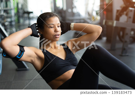 Focused young Black woman doing crunches on gym floor, hands behind head Focused young Black woman doing crunches on gym floor, hands behind head 139183473
