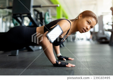 Focused Black woman doing push-ups on gym floor, wearing black sports bra 139183495