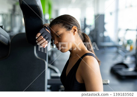 Exhausted young Black woman leaning head against gym machine with eyes closed Exhausted young Black woman leaning head against gym machine with eyes closed 139183502