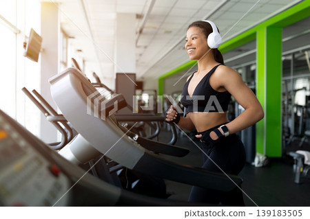 Smiling young Black woman jogging on treadmill in gym, wearing white headphones Smiling young Black woman jogging on treadmill in gym, wearing white headphones 139183505