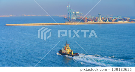 Manama, Bahrain - Jan 08 2024, Panoramic view of passing tugboat, a boat, in the background a commercial international port with cranes, pier, buildings and containers, in the daytime, Manama, Bahrain Manama, Bahrain - Jan 08 2024, Panoramic view of passing tugboat, a boat, in the background a commercial international port with cranes, pier, buildings and containers, in the daytime, Manama, Bahrain 139183635