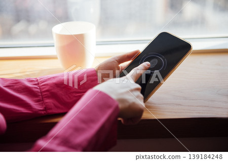 Female user in pink shirt interacts with smartphone on wooden windowsill, coffee cup visible in the background, focusing on fingerprint recognition feature Female user in pink shirt interacts with smartphone on wooden windowsill, coffee cup visible in the background, focusing on fingerprint recognition feature 139184248